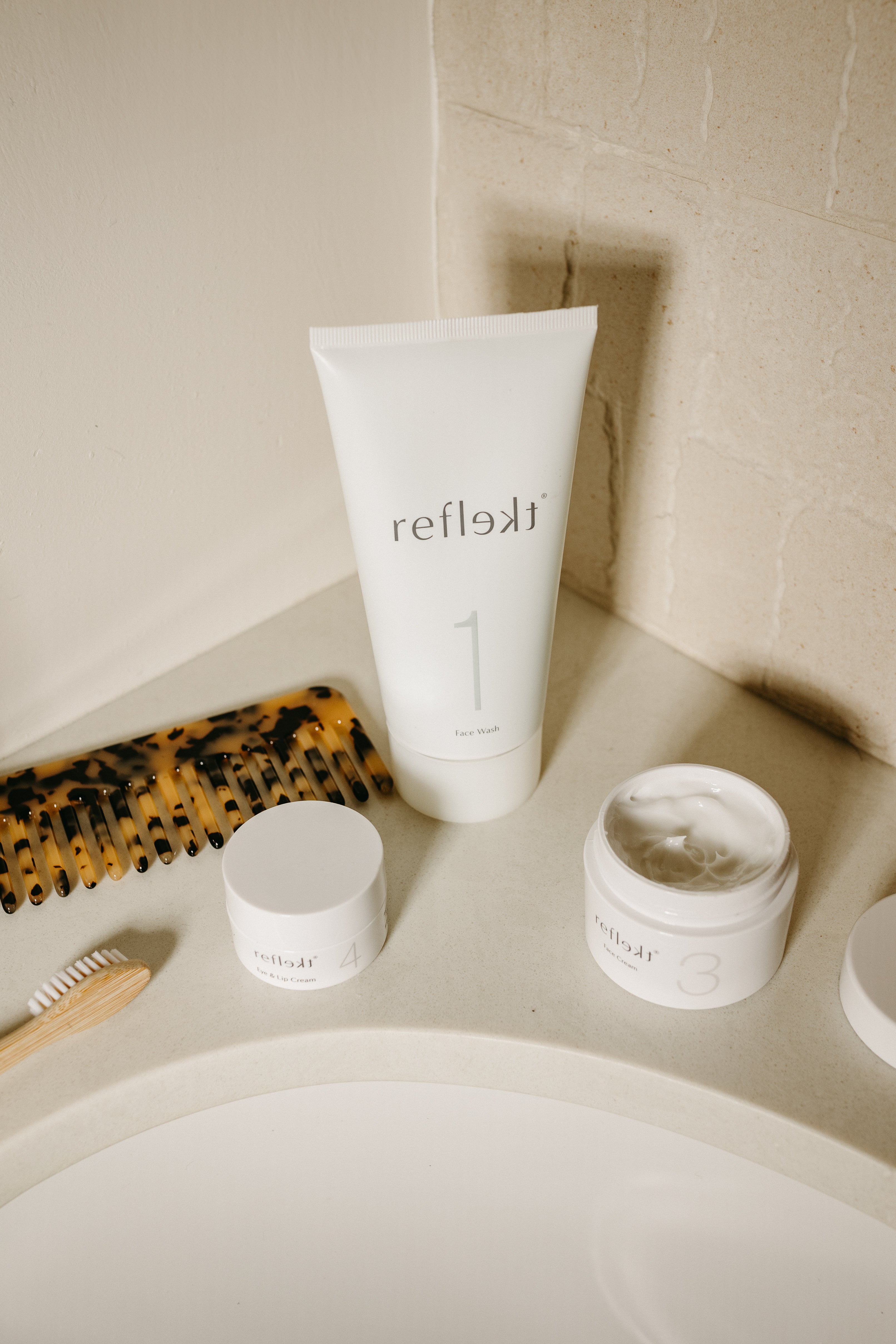 Reflekt skincare bottles on bathroom sink with comb and toothbrush blurred in the background